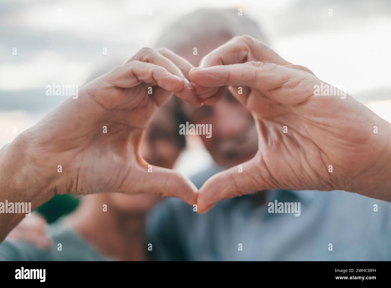 Close up portrait happy sincere middle aged elderly retired family couple making heart gesture with fingers, showing love or demonstrating sincere feelings together outdoors, looking at camera. Stock Photo