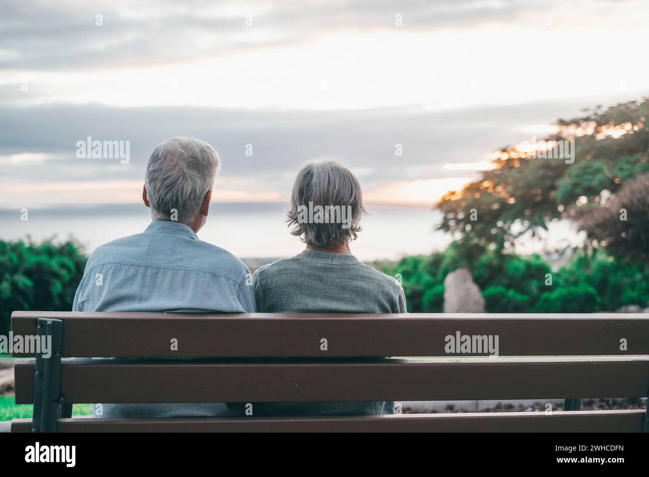 Head shot close up portrait happy grey haired middle aged woman with older husband, enjoying sitting on bench at park. Bonding loving old family couple embracing, looking sunset. Stock Photo