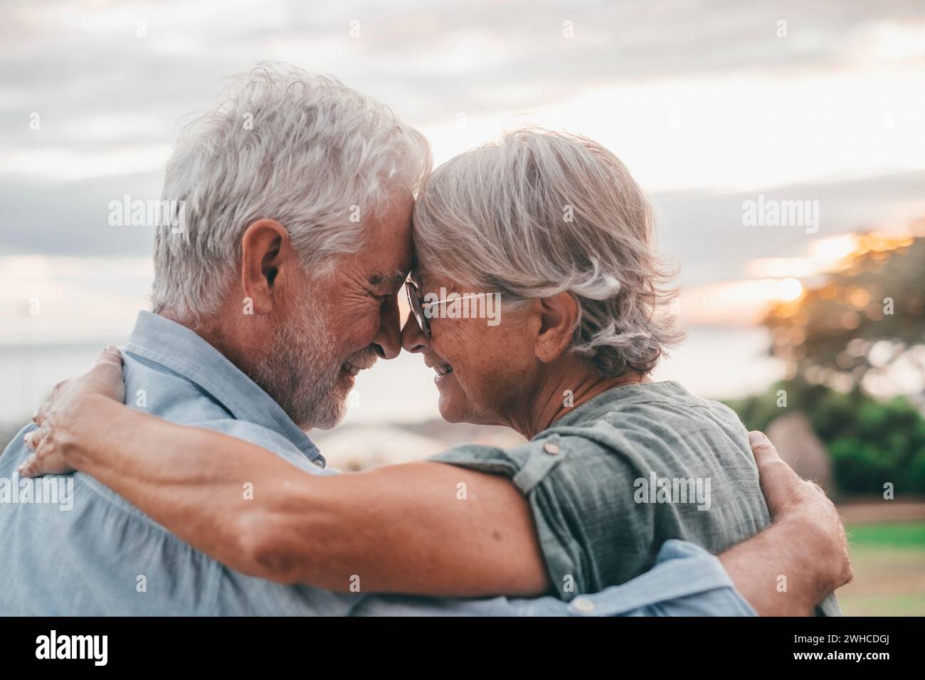Head shot close up portrait happy grey haired middle aged woman snuggling to smiling older husband, enjoying sitting on bench at park. Bonding loving old family couple embracing, looking sunset. Stock Photo