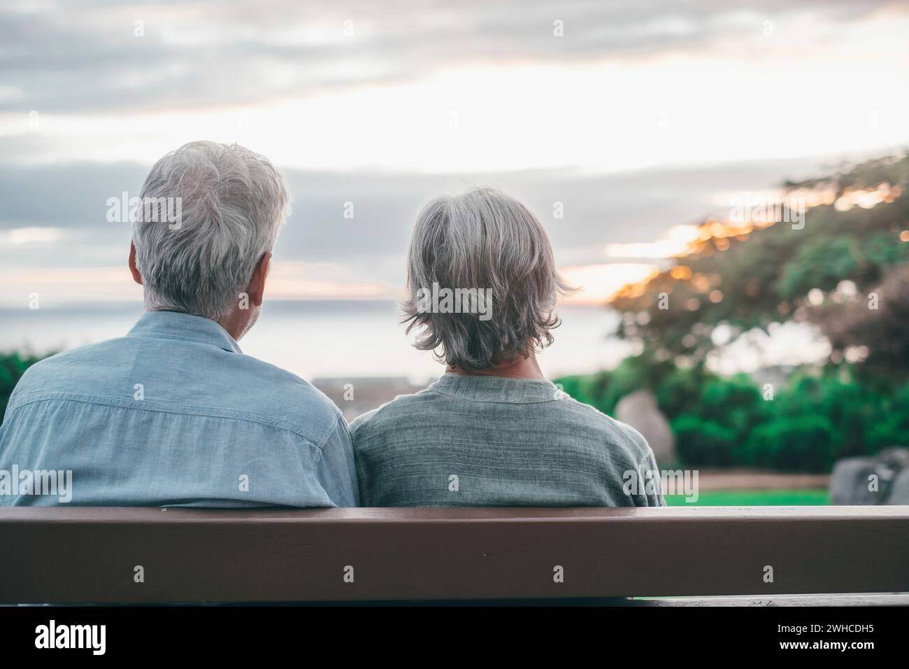 Head shot close up portrait happy grey haired middle aged woman with older husband, enjoying sitting on bench at park. Bonding loving old family couple embracing, looking sunset. Stock Photo