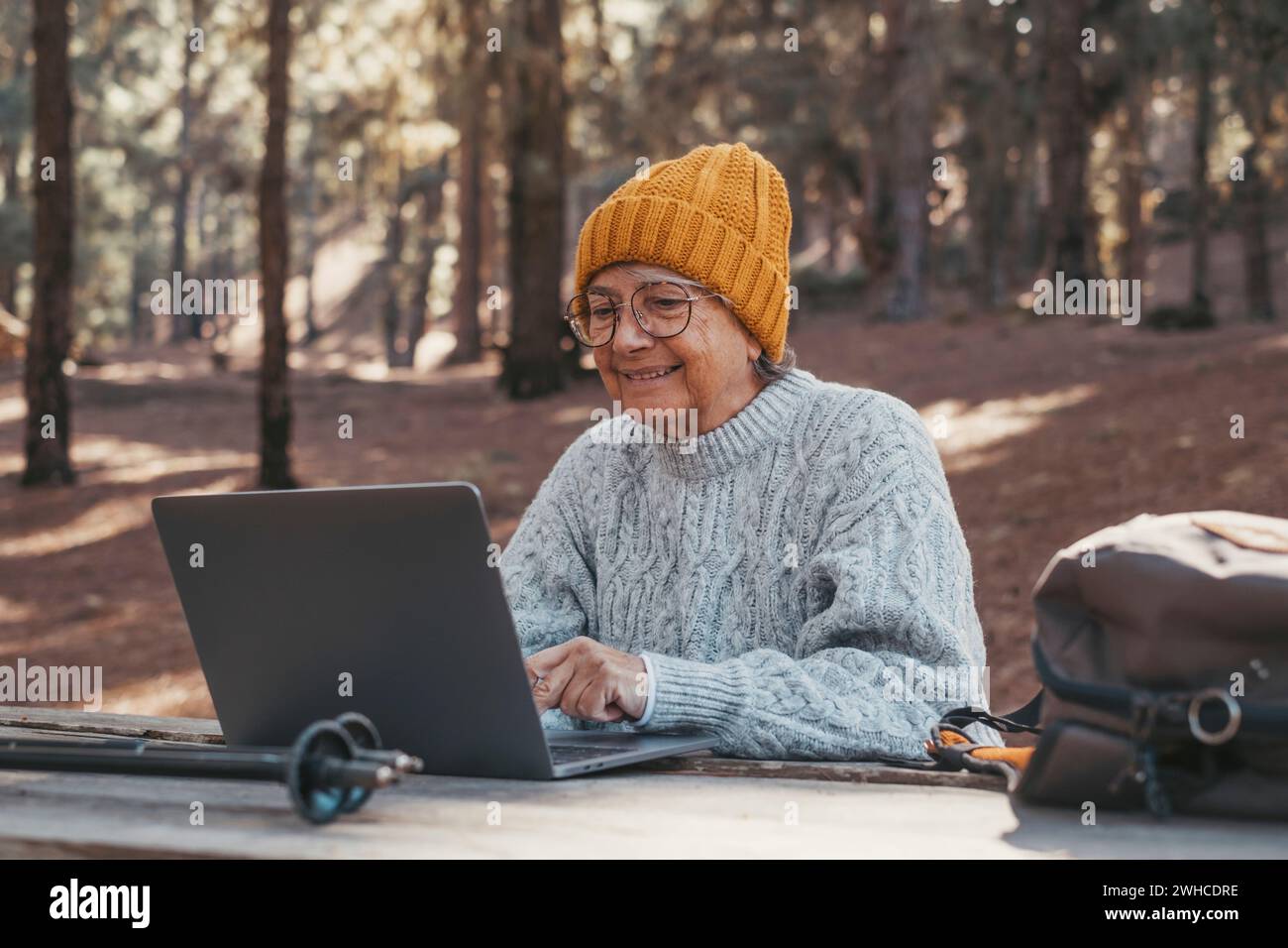 Portrait, close up of cute one of old middle age person using computer pc outdoors sitting at a wooden table in the forest of mountain in nature with trees around her. Stock Photo