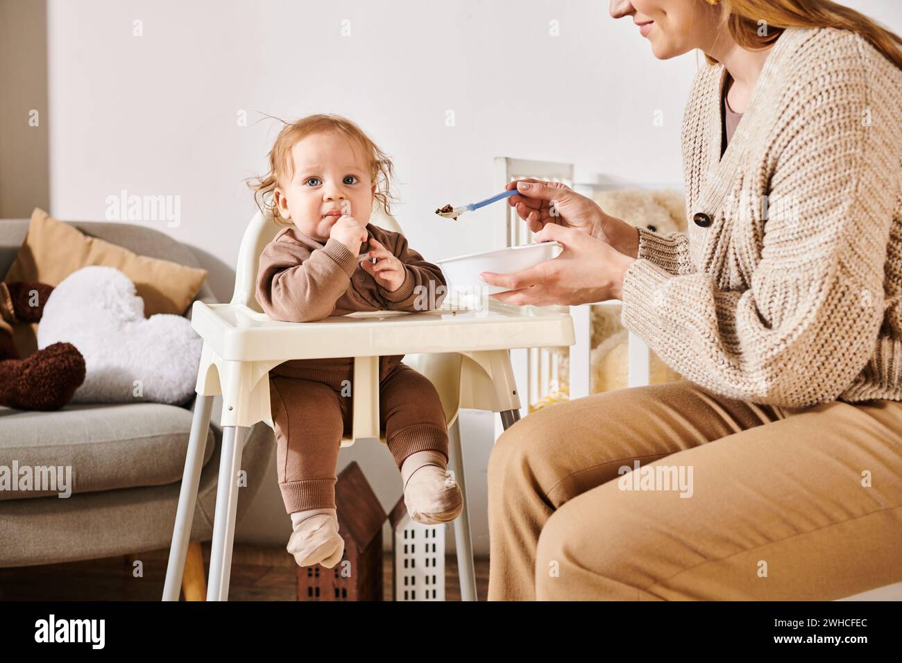 smiling woman feeding toddler boy with breakfast on baby chair in nursery room, blissful motherhood Stock Photo