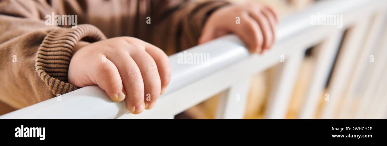 childhood, tiny hands of little kid standing in crib in nursery room at home, horizontal banner Stock Photo