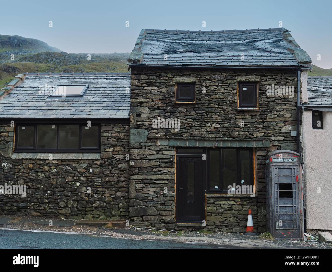 old British telephone kiosk painted grey, in a state of disrepair outside a traditional Lakeland stone building. Phone boxes are usually painted red. Stock Photo