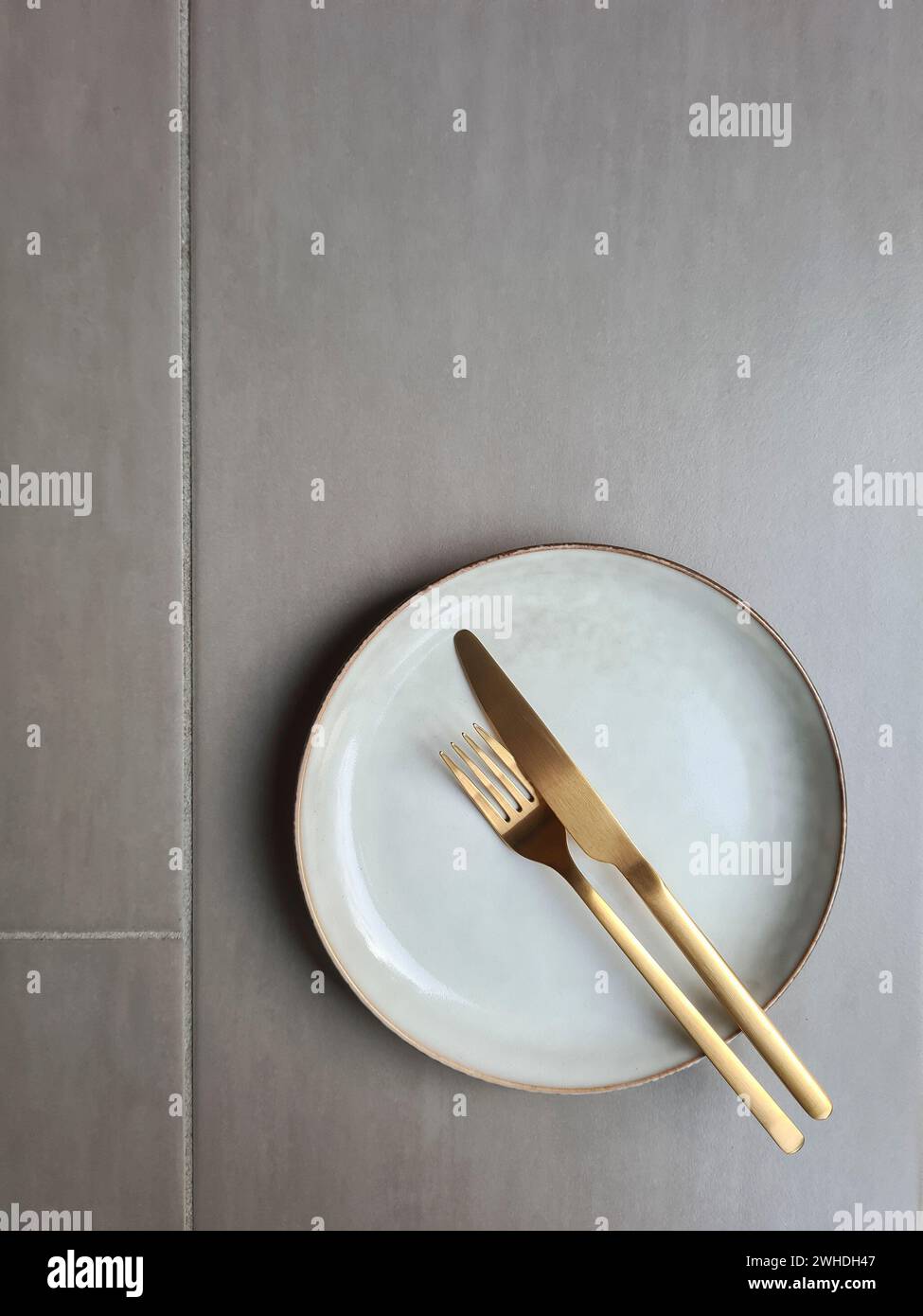 Natural-colored ceramic plate with brown rim and gold-colored knife with fork on a grey background, plate with cutlery Stock Photo