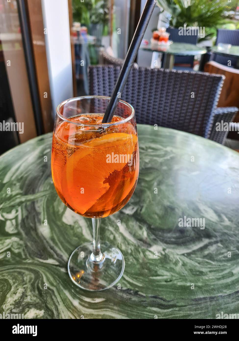 The alcoholic soft drink Aperol Spritz with a slice of orange in the glass and a black straw stands alone on a table outside the restaurant Stock Photo