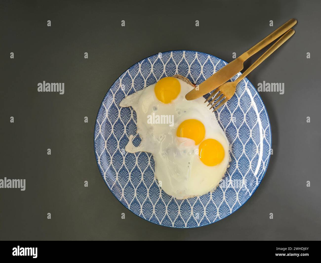 Three fried eggs on a blue and white plate with gold-colored cutlery, knife and fork Stock Photo