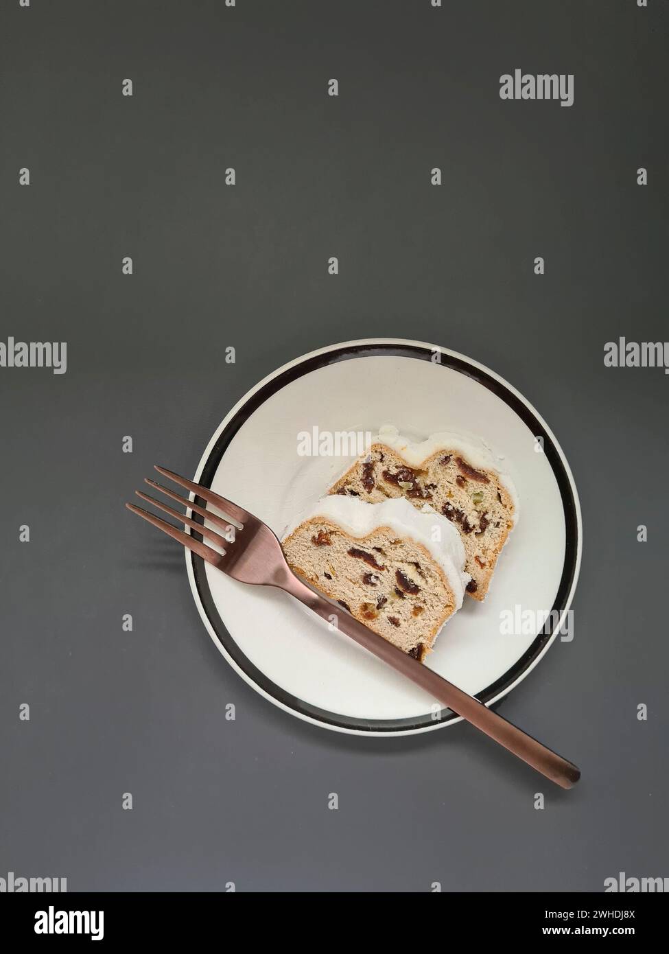 Two slices of Christmas cake with white powdered sugar on a white plate with a black rim and a copper-colored fork Stock Photo