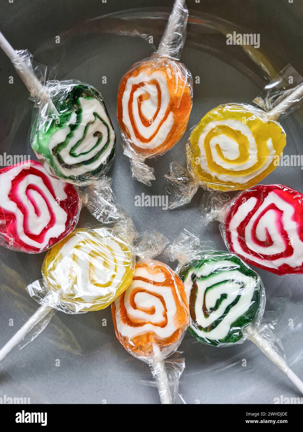 Eight colorful lollipops are still wrapped in plastic foil in a circle in a gray bowl Stock Photo