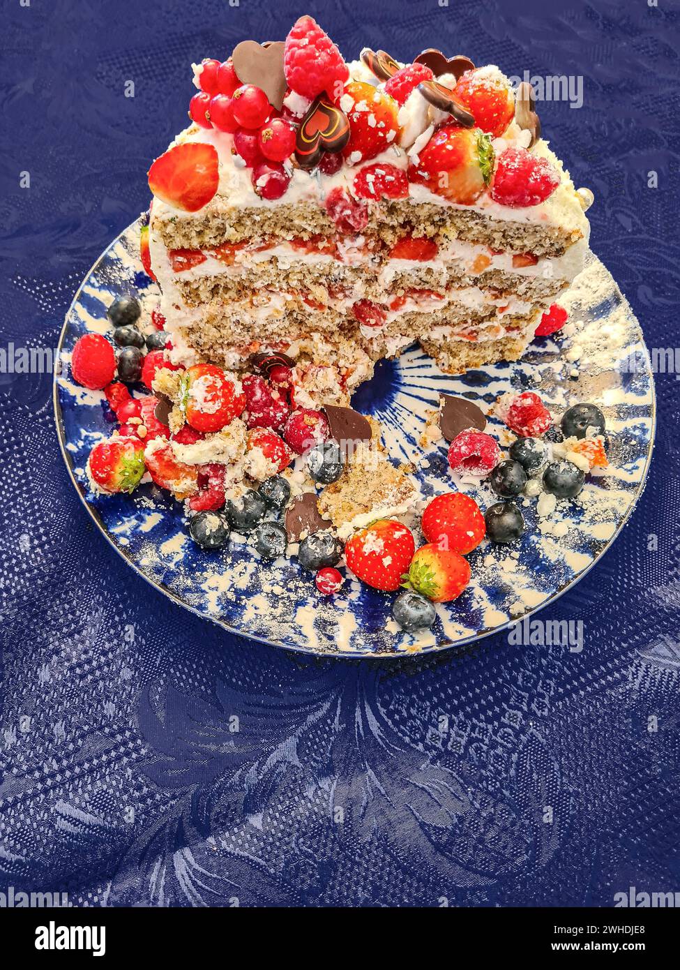 A festive cream cake with fruits such as strawberries, raspberries, currants and blueberries as decoration is cut and shows the cross-section of the cake and its individual layers Stock Photo