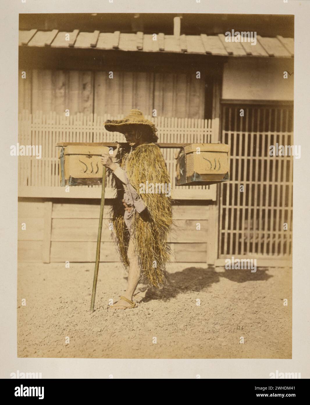 Japanese Porter wearing protective outer garments made of straw..  Photograph shows a porter, full-length, standing, facing left, wearing a hat and outer garments made of straw for protection against rain and snow over his shoulders and around his waist, carrying shoulder pole with boxes attached over his right shoulder, and holding staff in left hand....Japan  Hand-coloured with watercolour Photographic Print circa 1870s Stock Photo