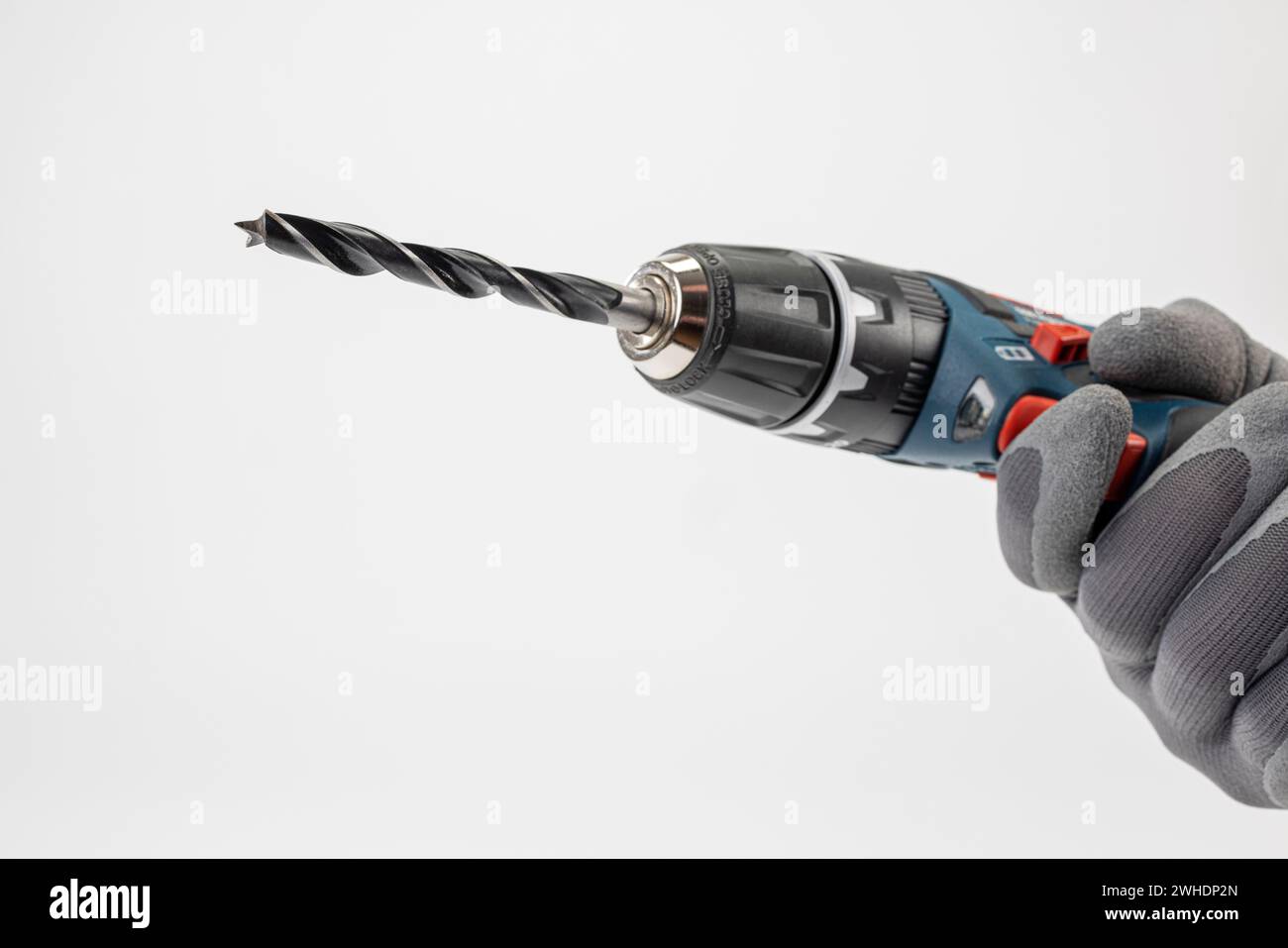 Man's hand with work glove holding Bosch cordless drill, with inserted wood drill, white background, Stock Photo