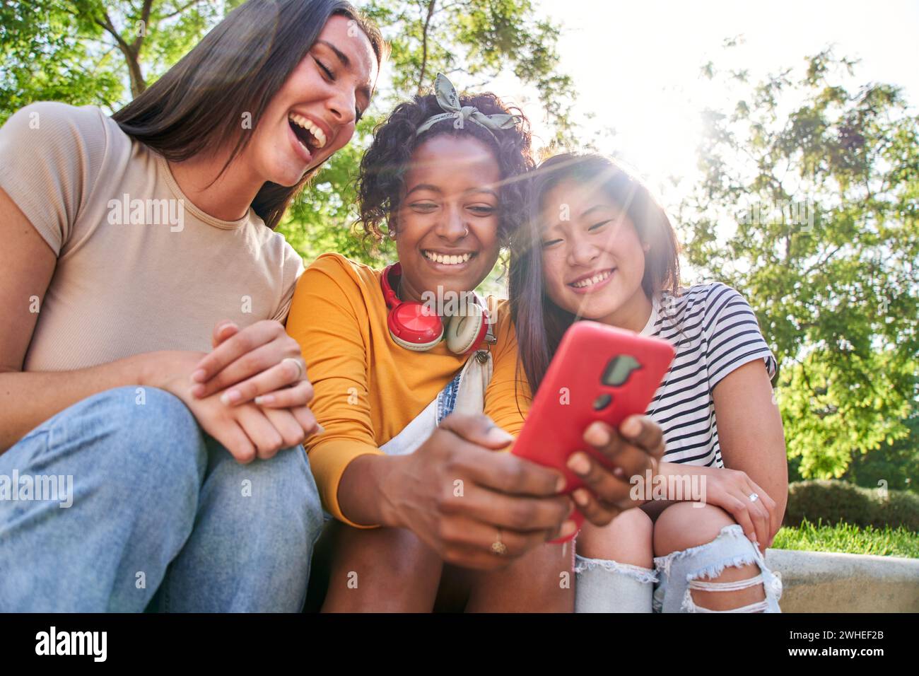 Group joyful friends using mobile sitting outdoor. Three laughing multiracial women scrolling phone Stock Photo