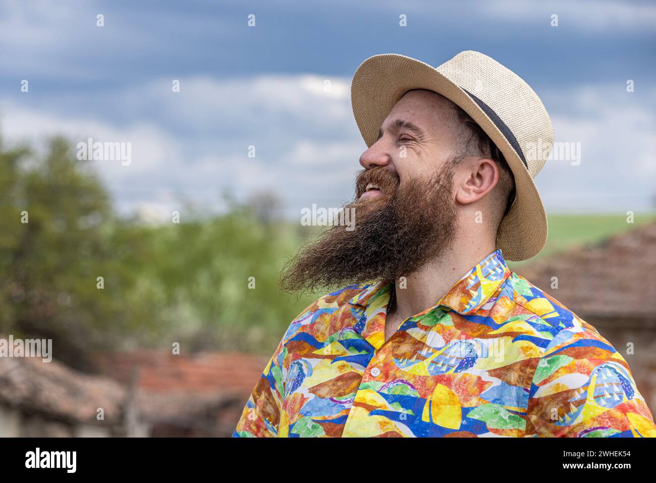 Young bearded man in fedora hat standing smiling happily. Close-up portrait of a bearded young man wearing casual clothes looking at the camera with a Stock Photo