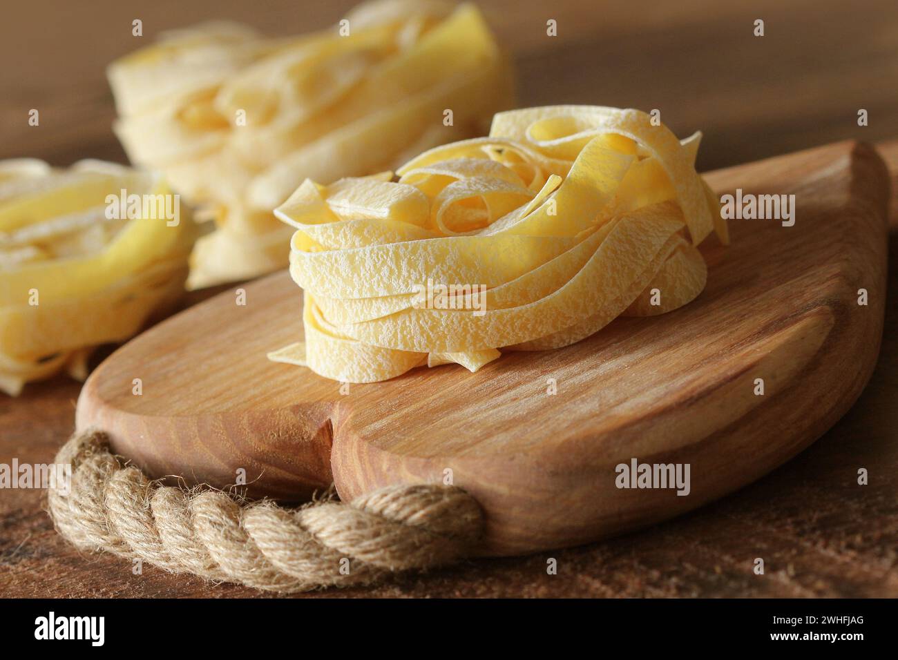 Nest of pasta tagliatelle on wooden cutting board aon rustic background. Selective focus Stock Photo