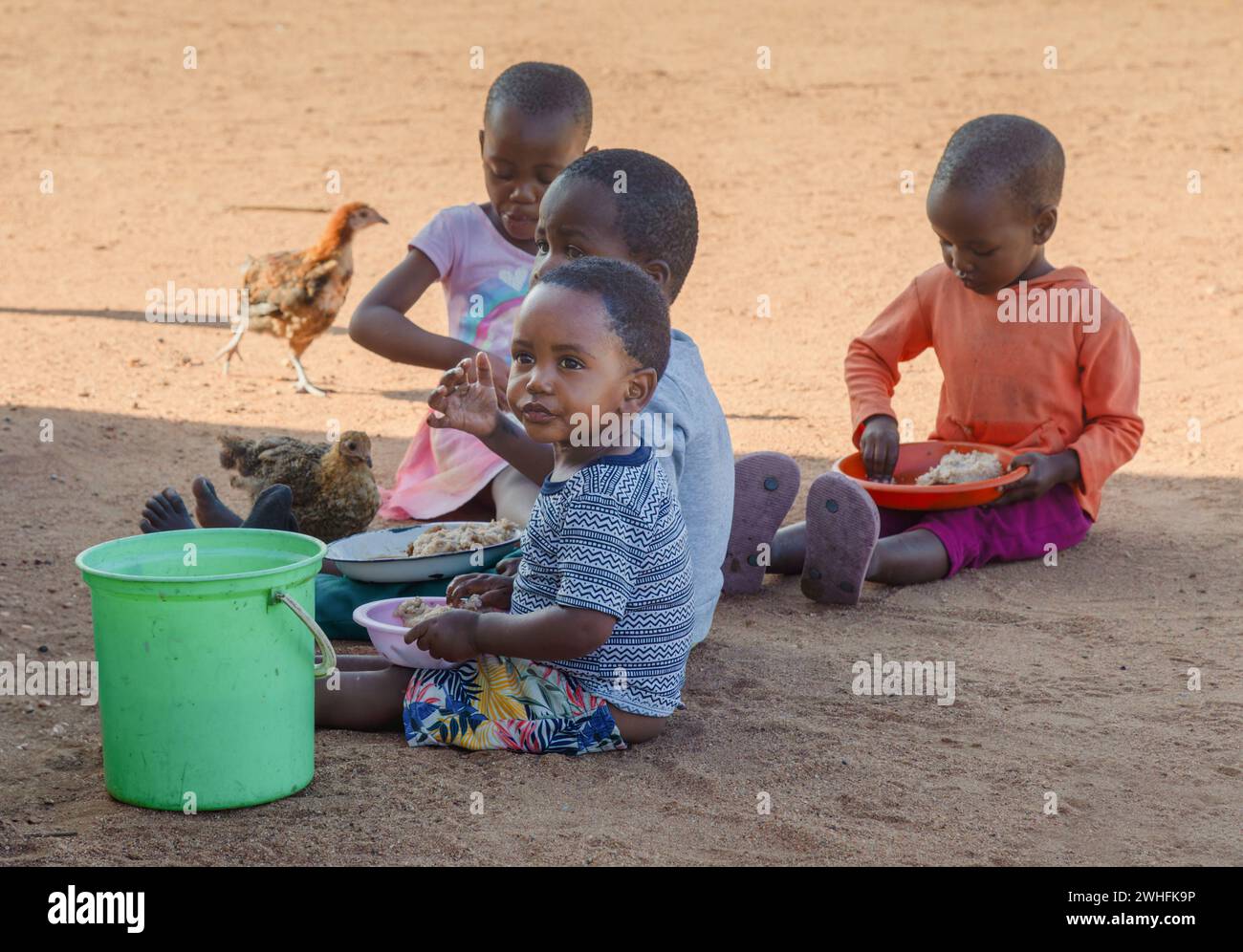 hungry village african children in the yard , sited on the ground eating staple food from plastic plates Stock Photo