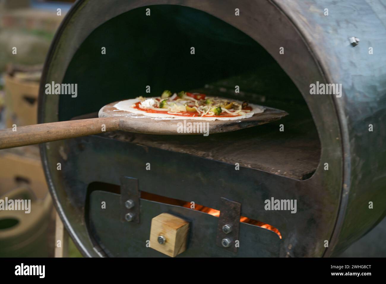 Fresh pizza on a wooden slide is pushed into a self-made recycling oven built from an old metal barrel, creative upcycling conce Stock Photo