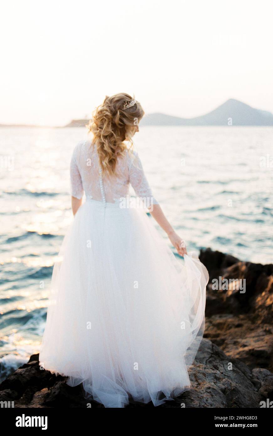 Bride holds the hem of a layered dress on the rocks by the sea. Back view Stock Photo