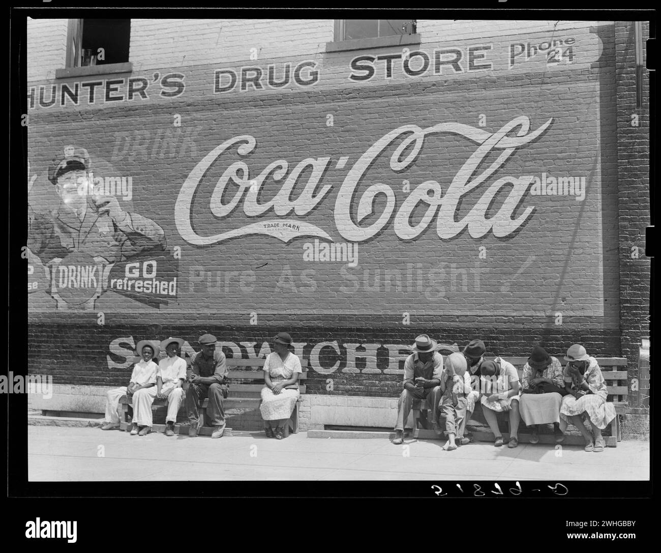 General scene, main street. Greensboro, Georgia, USA, circa 1930s.  People sitting on street bench in afternoon sun under a large Coca-Cola advertising painted on brick wall of Hunter's Drug Store Stock Photo