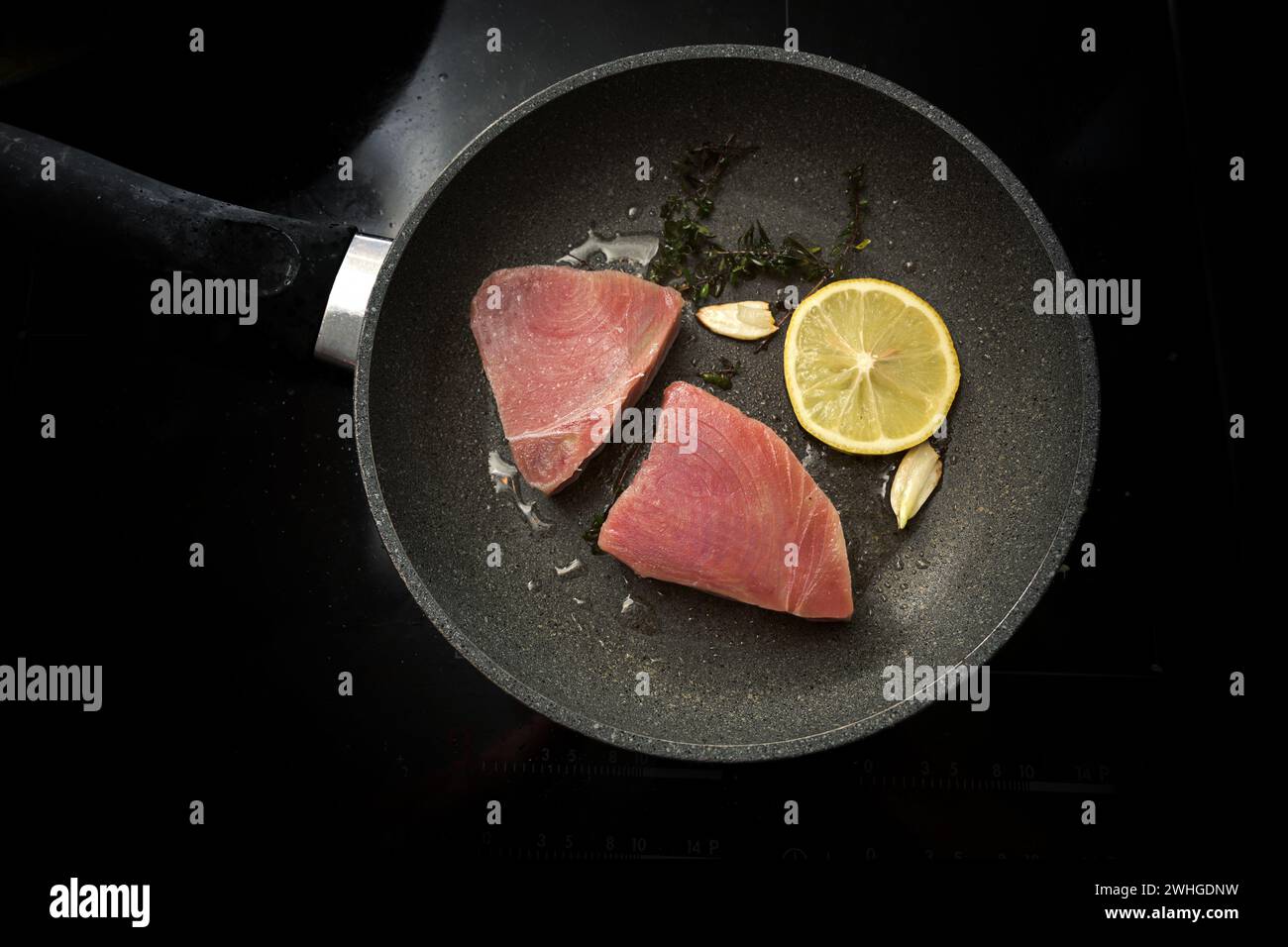 Fresh raw tuna steaks with lemon, garlic and thyme herb in a frying pan on a back stove, copy space, high angle view from direct Stock Photo