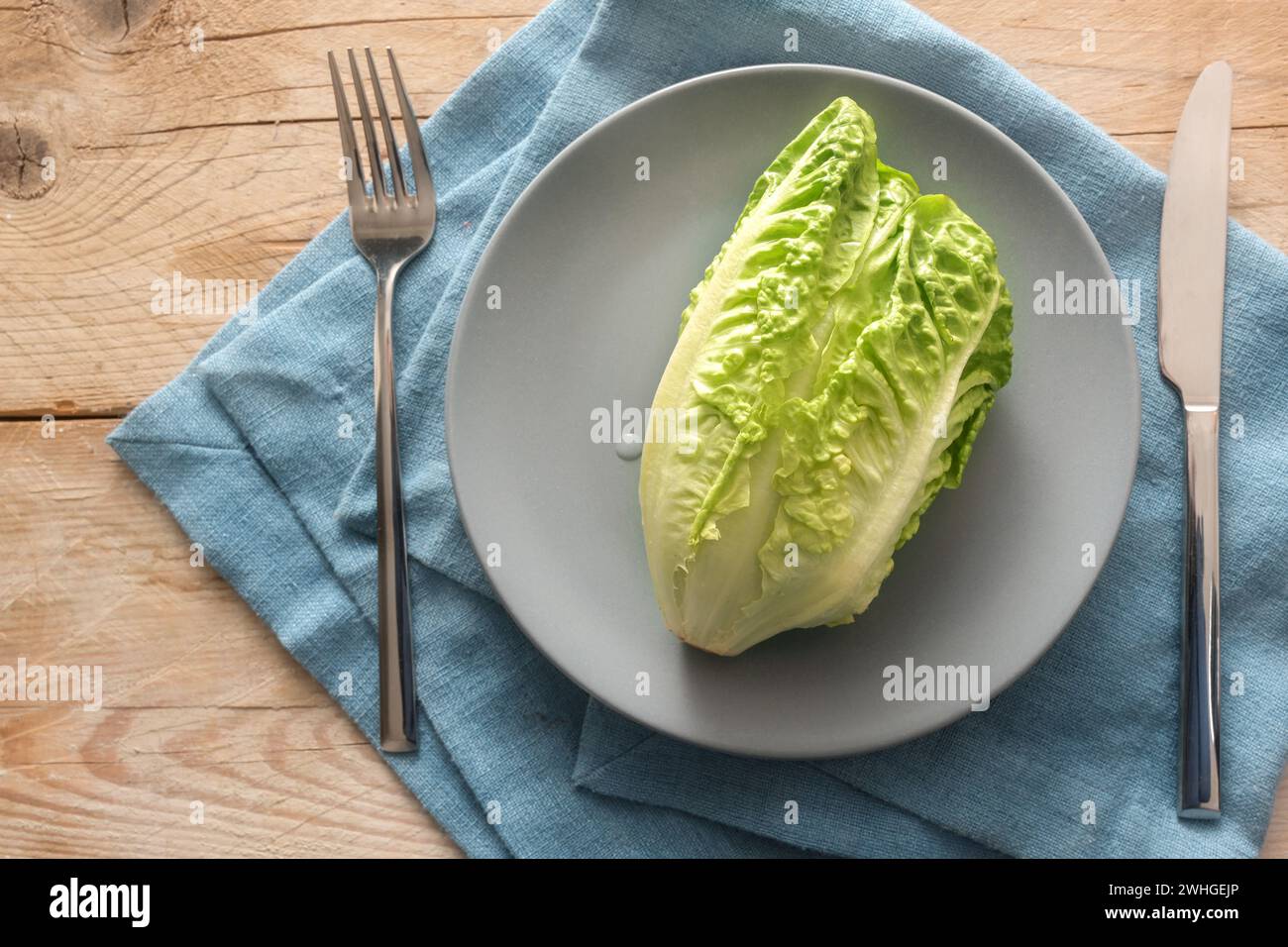 Fresh green lettuce or baby cos on a gray plate with cutlery on a blue napkin and rustic wooden planks, concept for a healthy ve Stock Photo