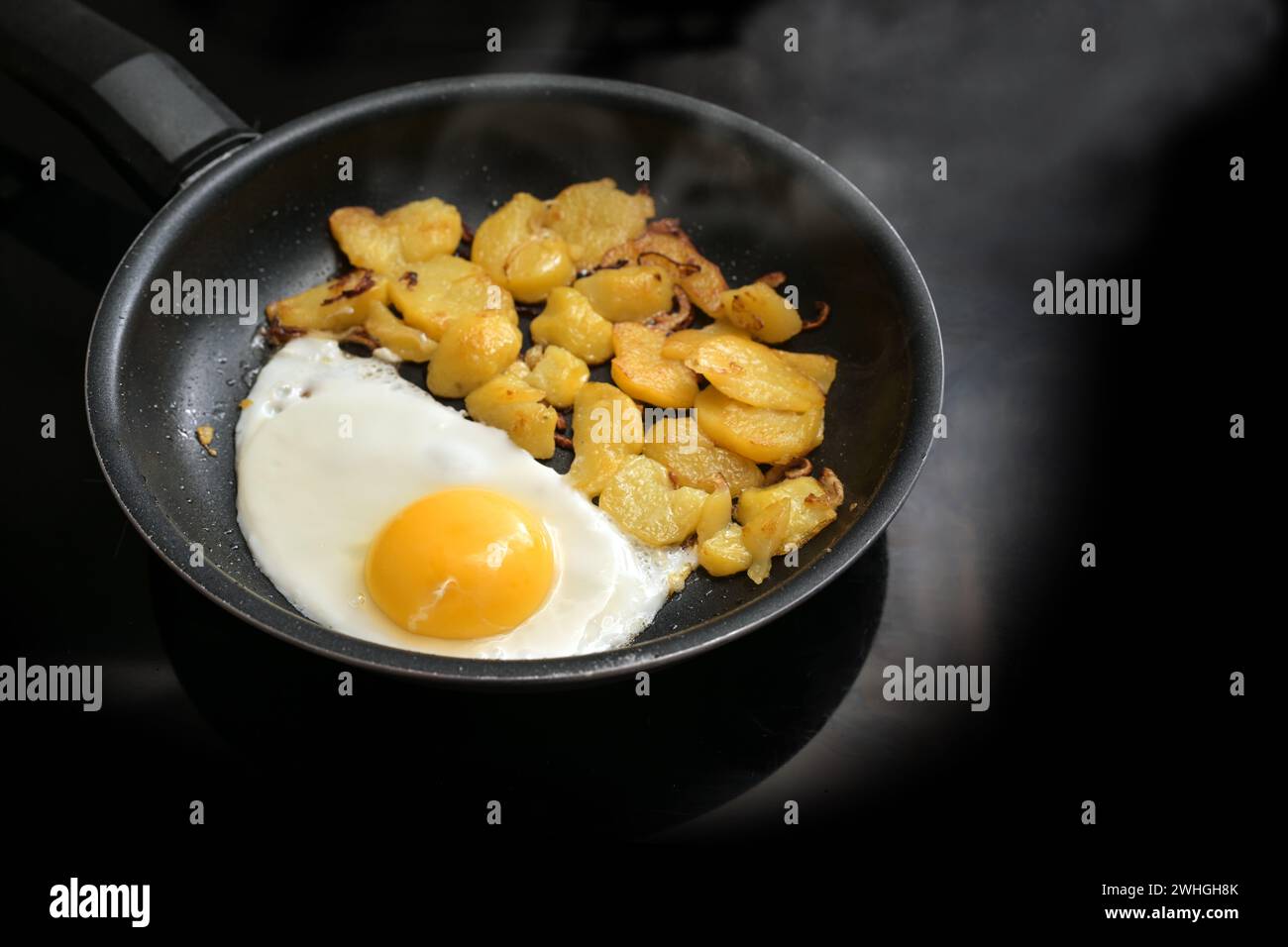 Fried egg and roast potatoes in a black frying pan on the stove in the kitchen, simple cooking, copy space, selected focus Stock Photo