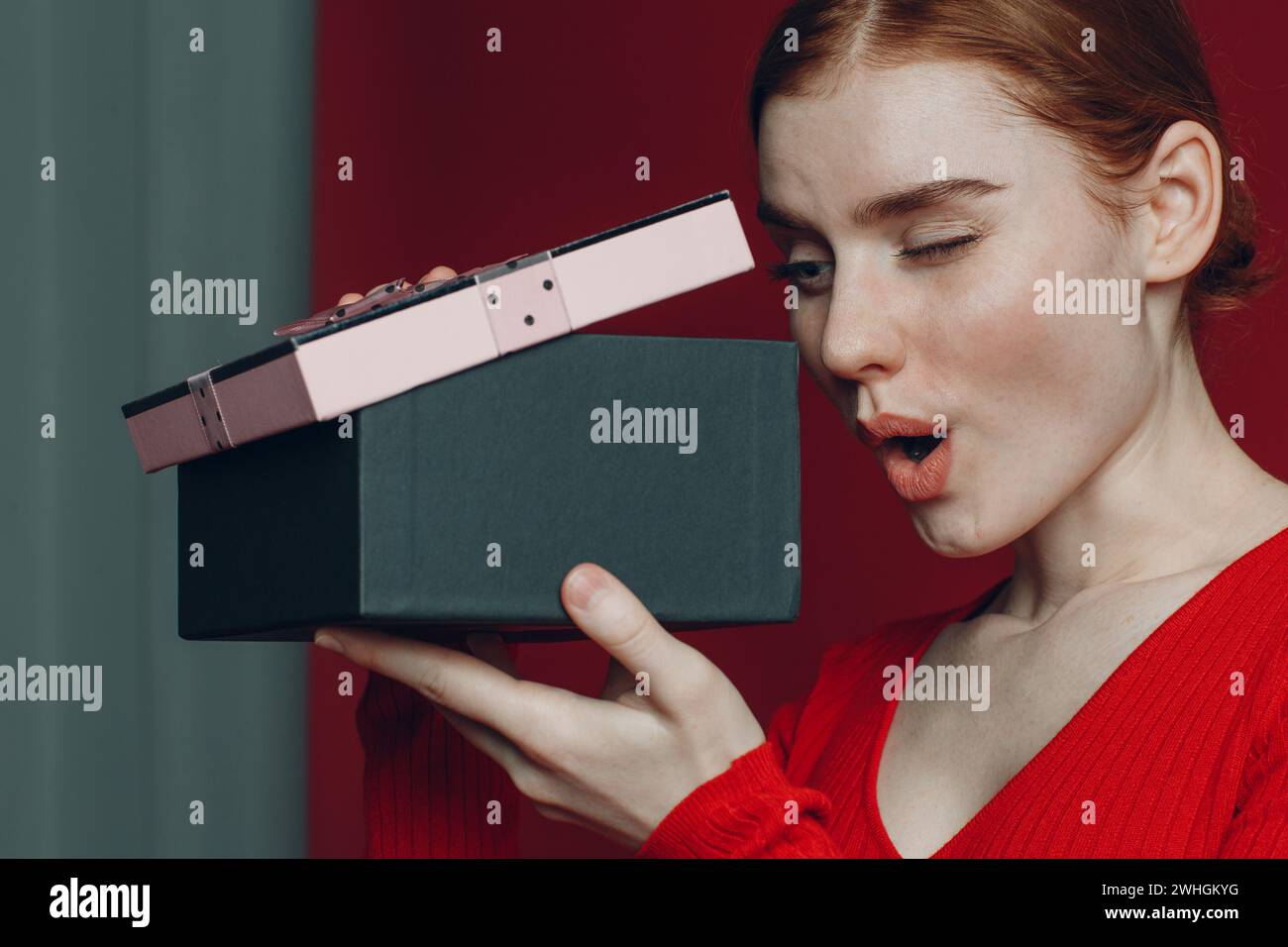 Red hair woman looks to pink gift box with ribbon. Studio shot portrait red background Stock Photo