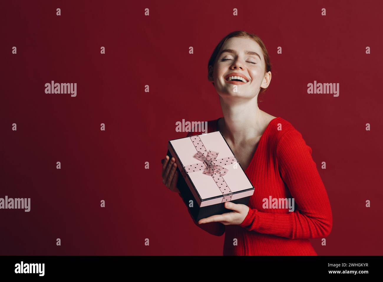 Red hair woman holding pink gift box with ribbon. Studio shot portrait red background Stock Photo