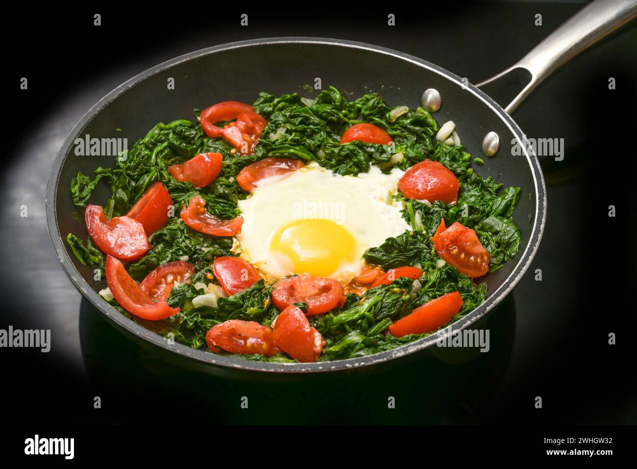 Fried egg with spinach and tomatoes in a frying pan on a black cooker top, preparing a healthy meal for breakfast or lunch, copy Stock Photo