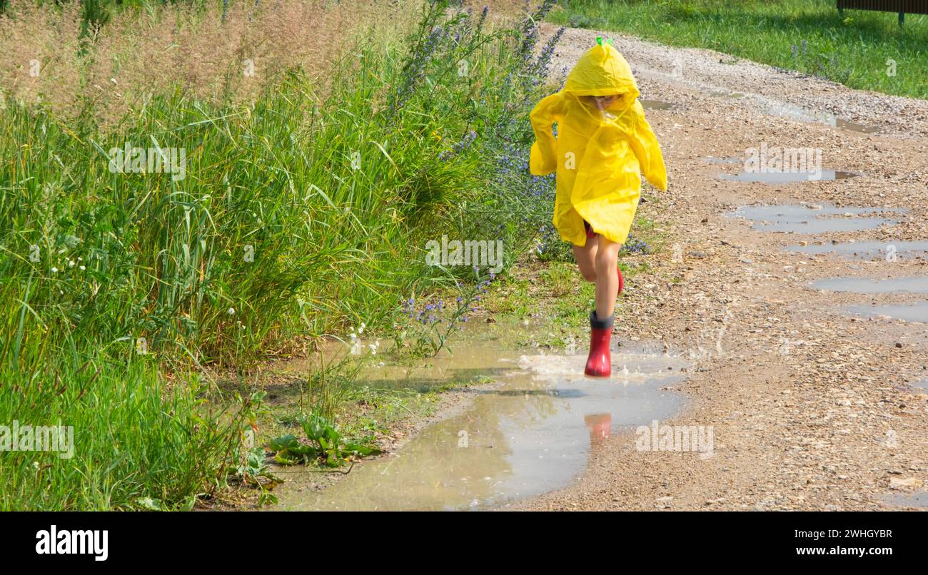 A girl in red rubber boots and a yellow raincoat runs through puddles ...