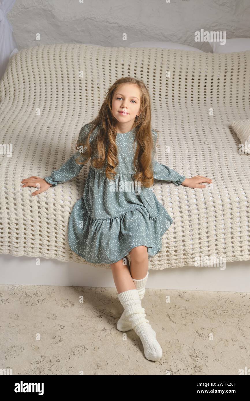 Cheerful young girl in pale mint dress sits on white knitted blanket spreads her arms out to the sides Stock Photo