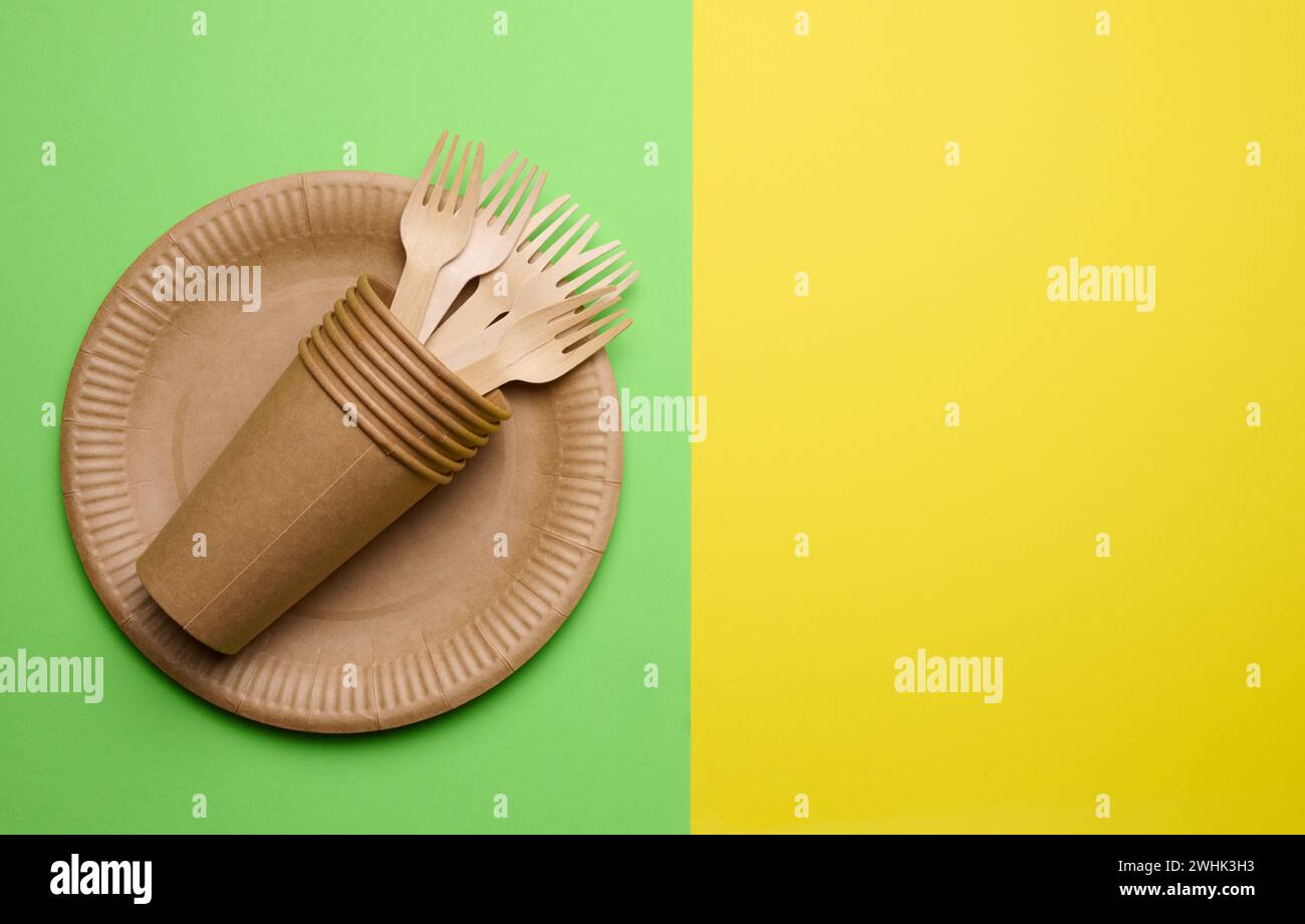 Brown paper plate, stack cups and wooden fork, disposable tableware on green background, top view. Copy space Stock Photo