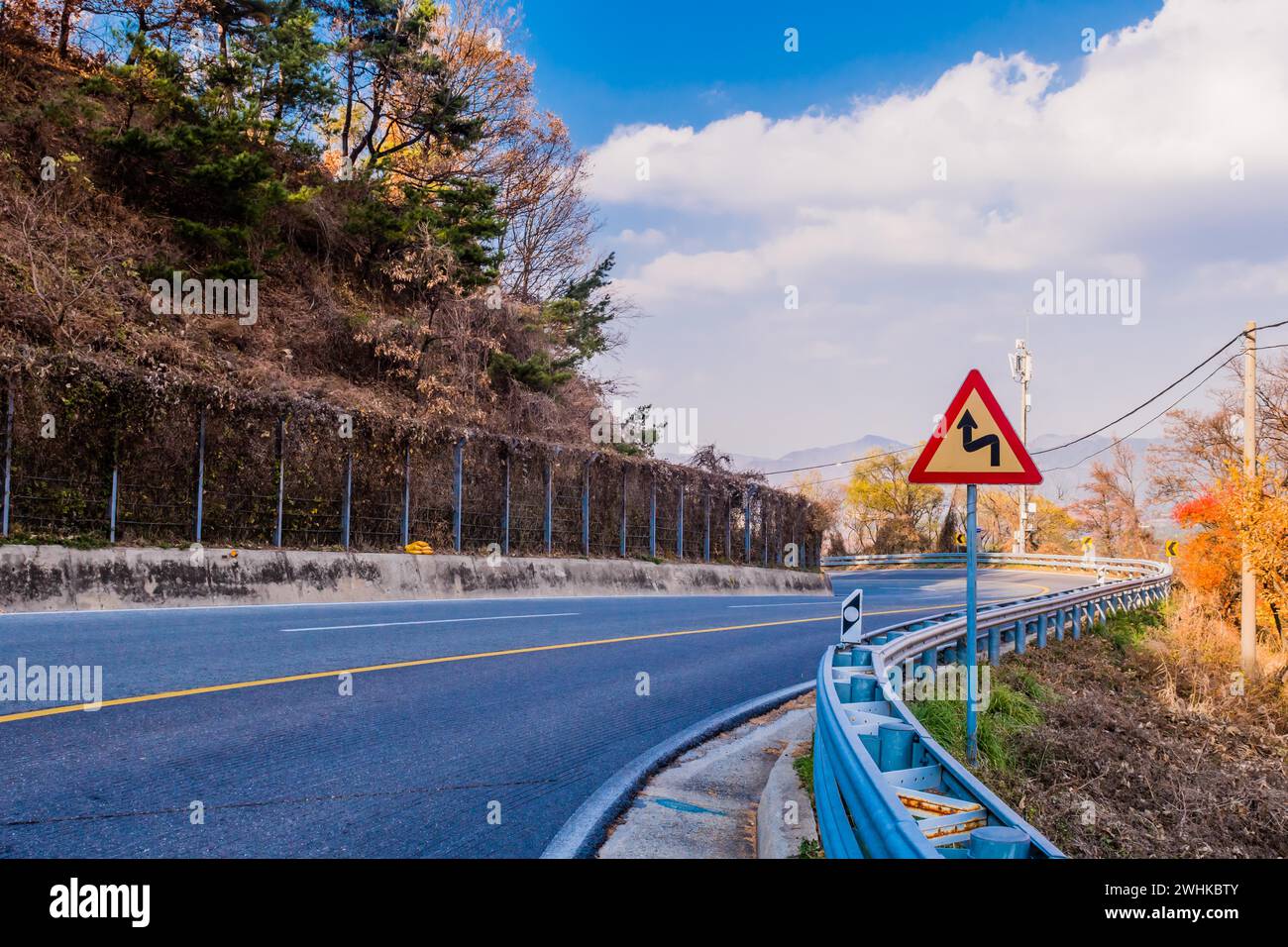 Warning sign next to guardrail on curvy mountain road with cloudy sky in background in South Korea Stock Photo