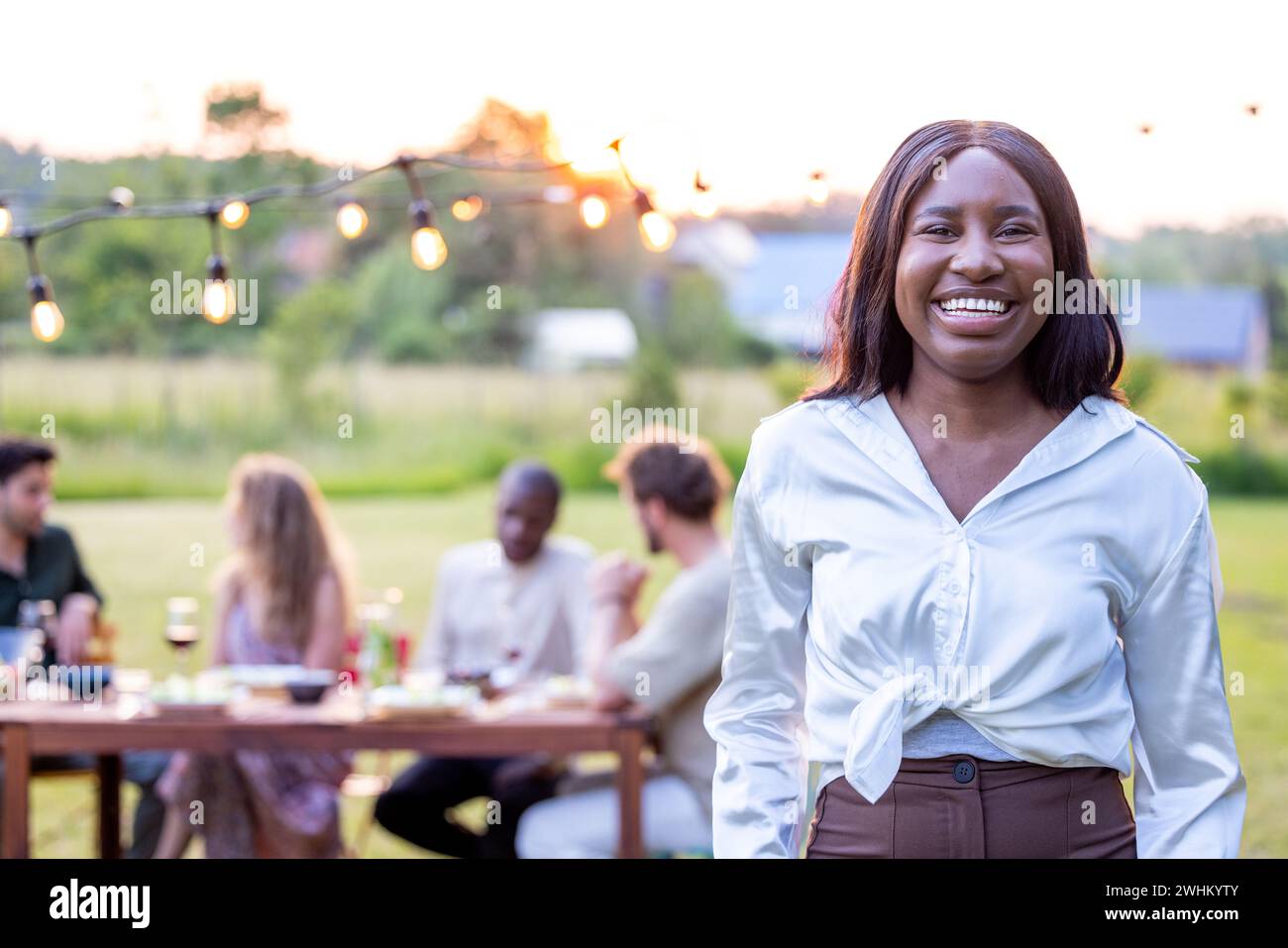 Beautiful young black woman with long straight hair in a white blouse with a group of friends in the background having an outdoo Stock Photo