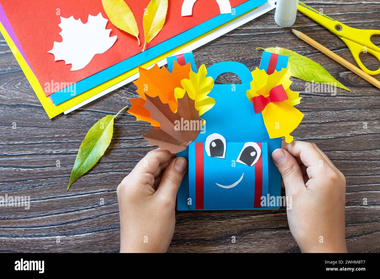 Child is holding Greeting card paper backpack on a wooden table. Handmade. Project of children's creativity, handicrafts, crafts Stock Photo