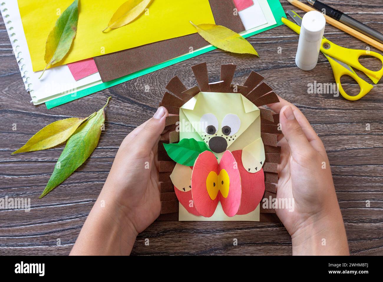 Child is holding backpack of paper greeting card and a postcard hedgehog with an apple on a wooden table. Handmade. Project of c Stock Photo