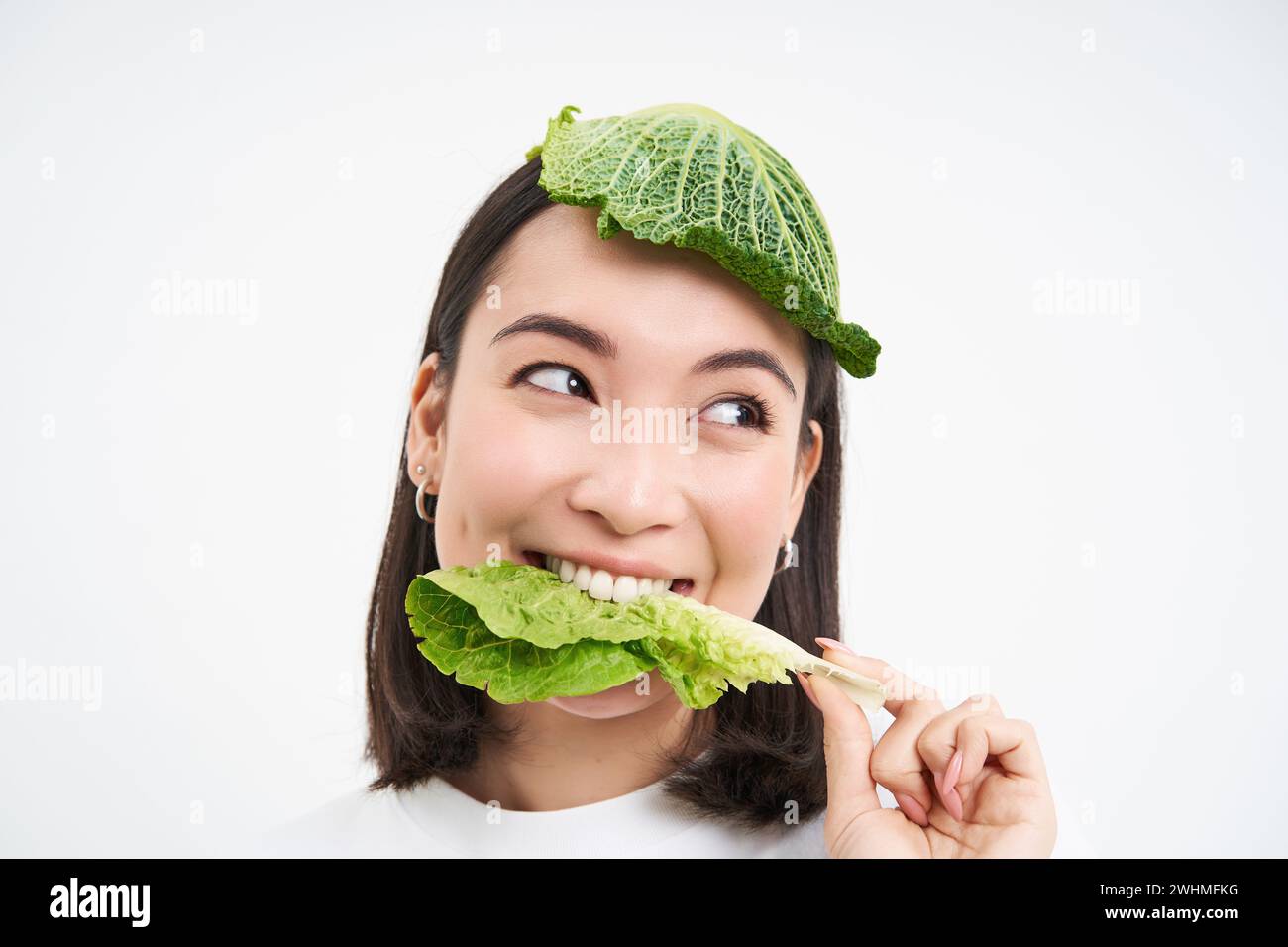Beautiful asian girl with lettuce on head, smiling and eating cabbage