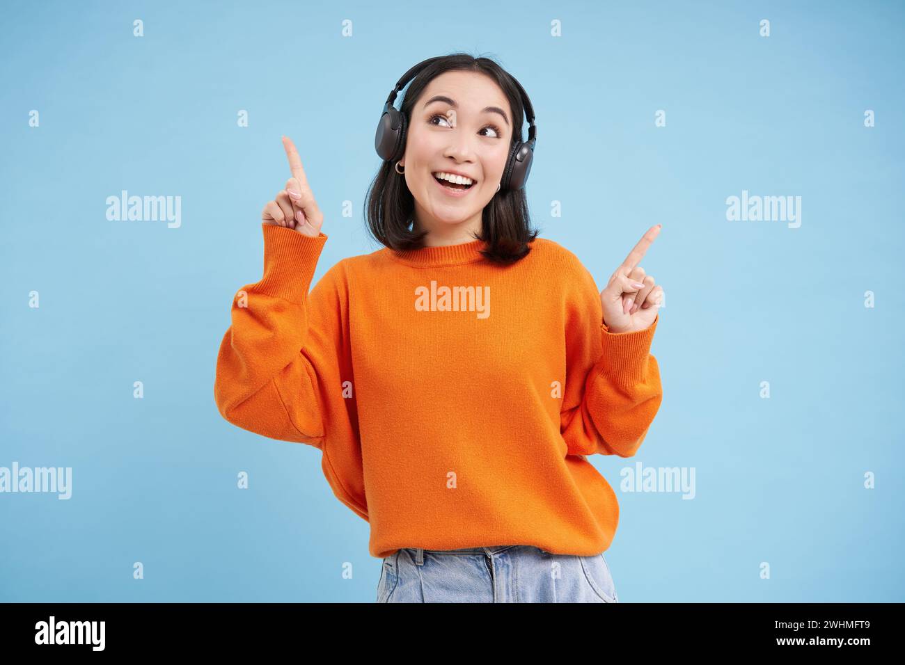 Happy Chinese woman in headphones, listens music, enjoys favourite song in her playlist, stands over blue background Stock Photo