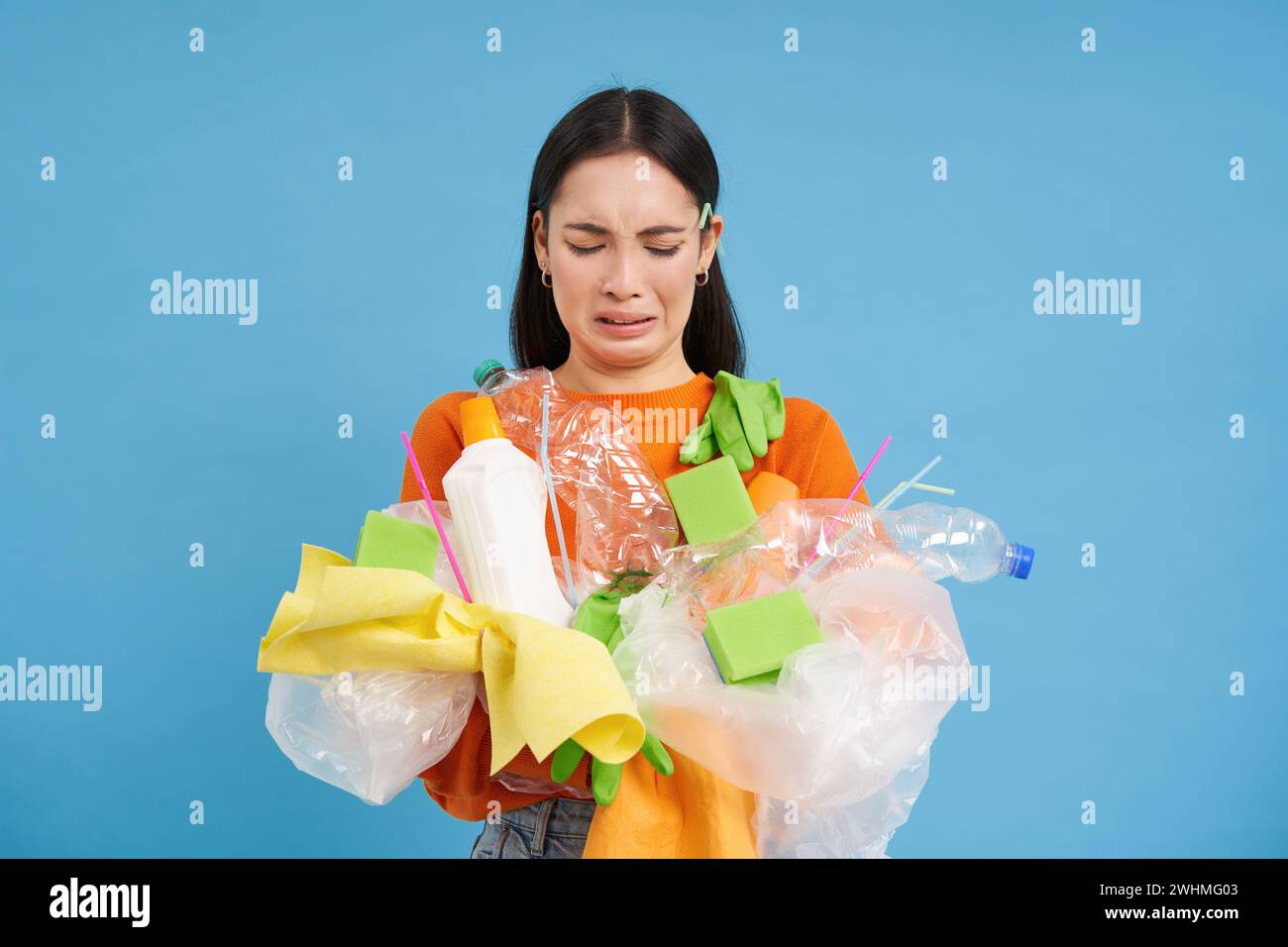 Frowning asian woman with plastic garbage in hands, complaining at non recycling waste, standing with litter over blue backgroun Stock Photo