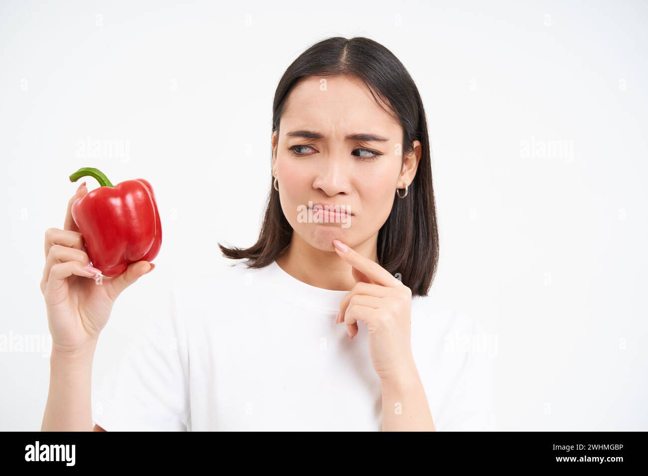 Close up portrait of pensive korean woman, looks at red pepper with thinking face, isolated on white background Stock Photo