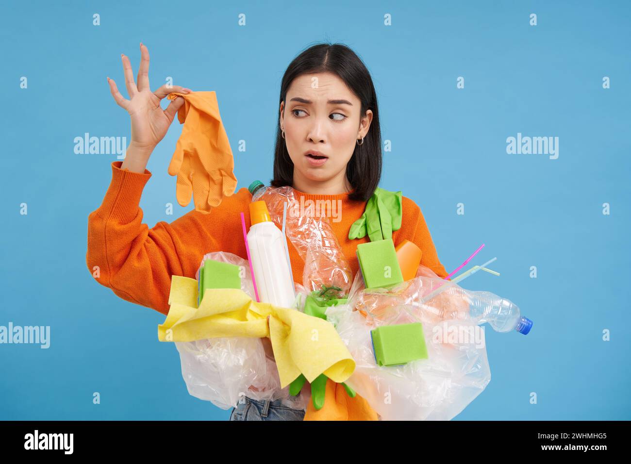 Young woman holds plastic garbage, looks at glove with shocked face, stinky waste, sorting litter for recycling, blue background Stock Photo