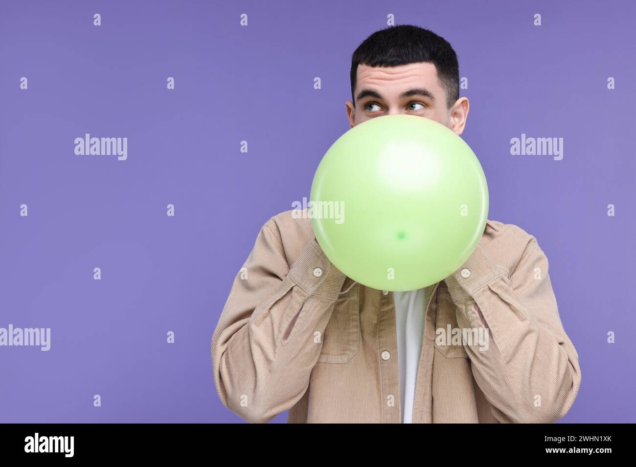 Young man inflating light green balloon on purple background. Space for ...
