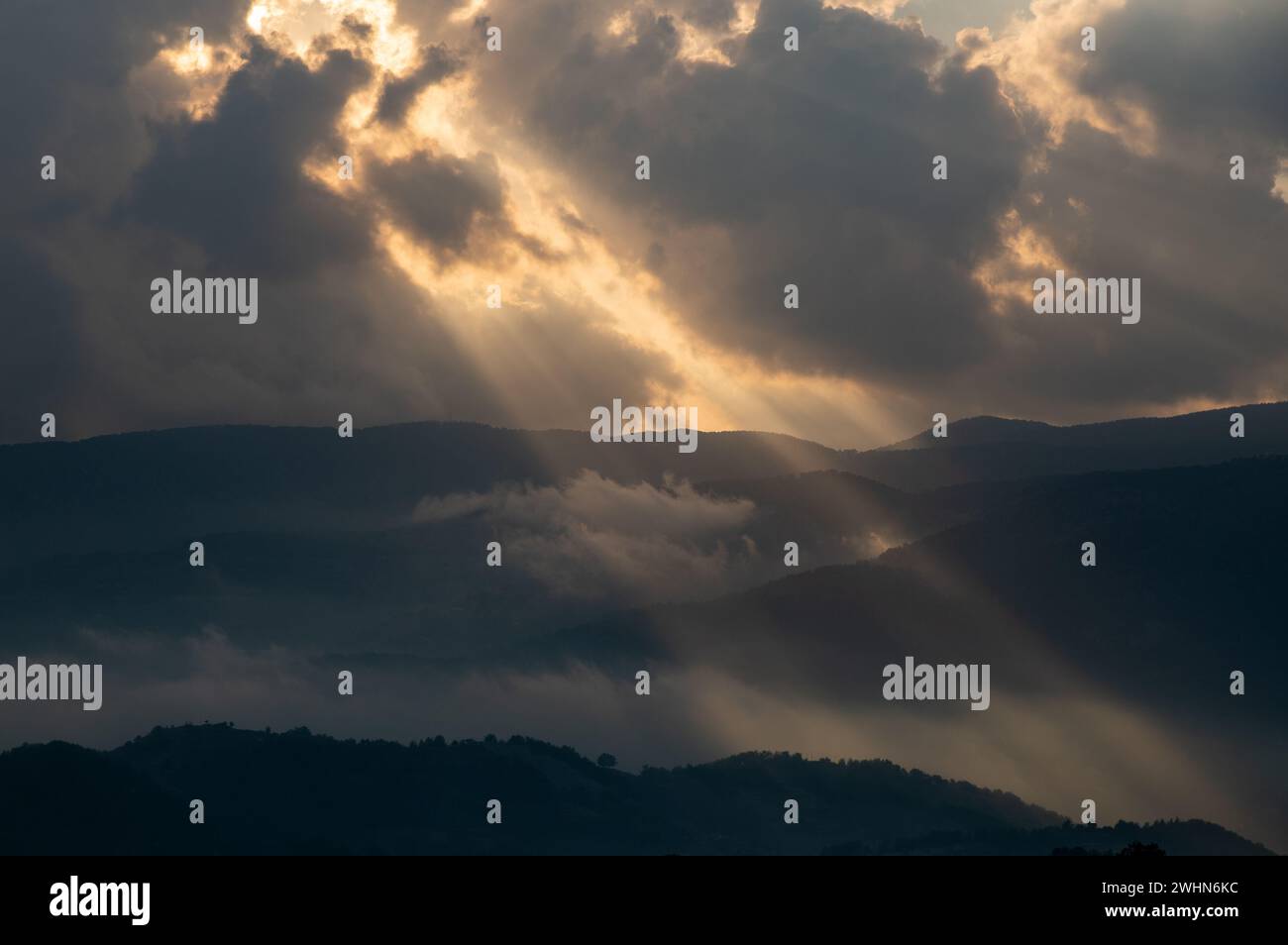 Bright sun rays light shining through dark clouds over mountain at sunset. Dramatic sky in winter Stock Photo