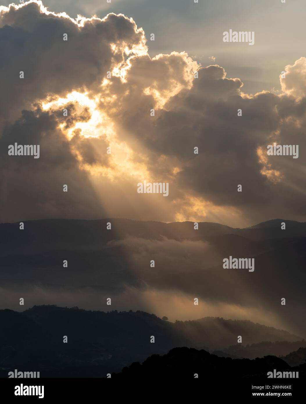 Bright sun rays light shining through dark clouds over mountain at sunset. Dramatic sky in winter Stock Photo