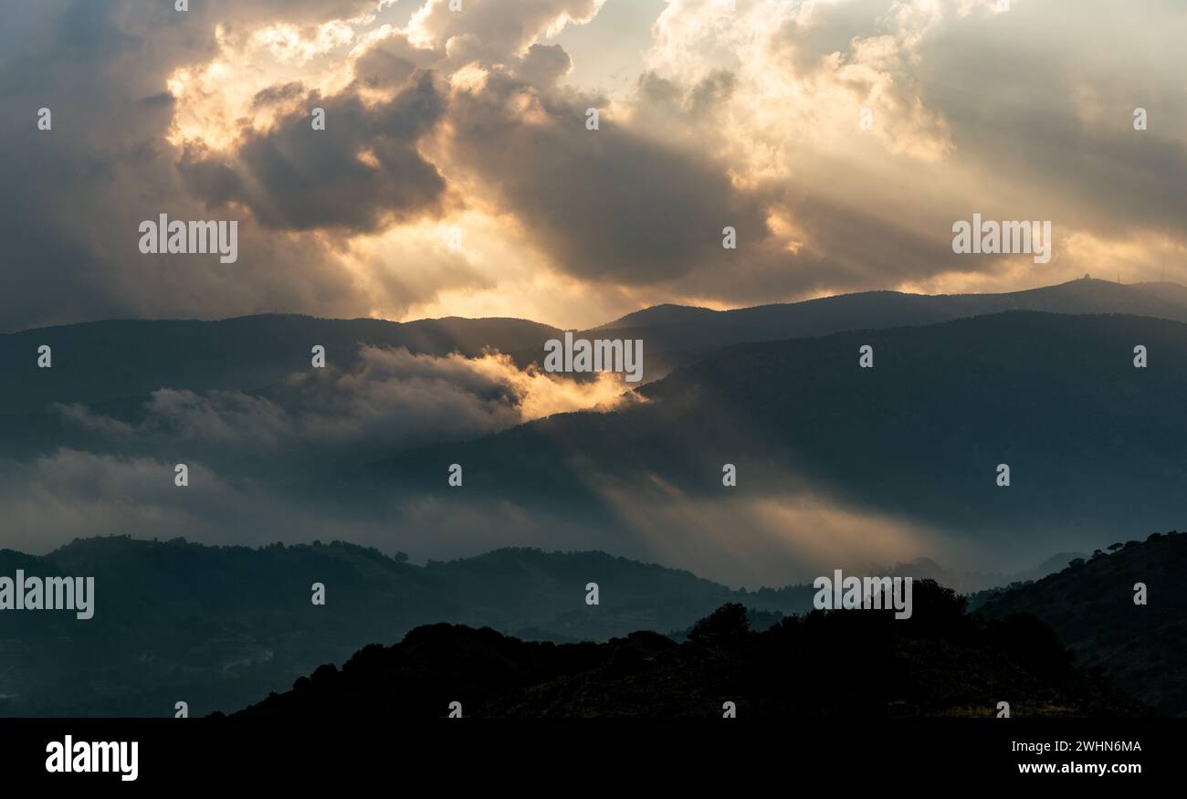 Bright sun rays light shining through dark clouds over mountain at sunset. Dramatic sky in winter Stock Photo