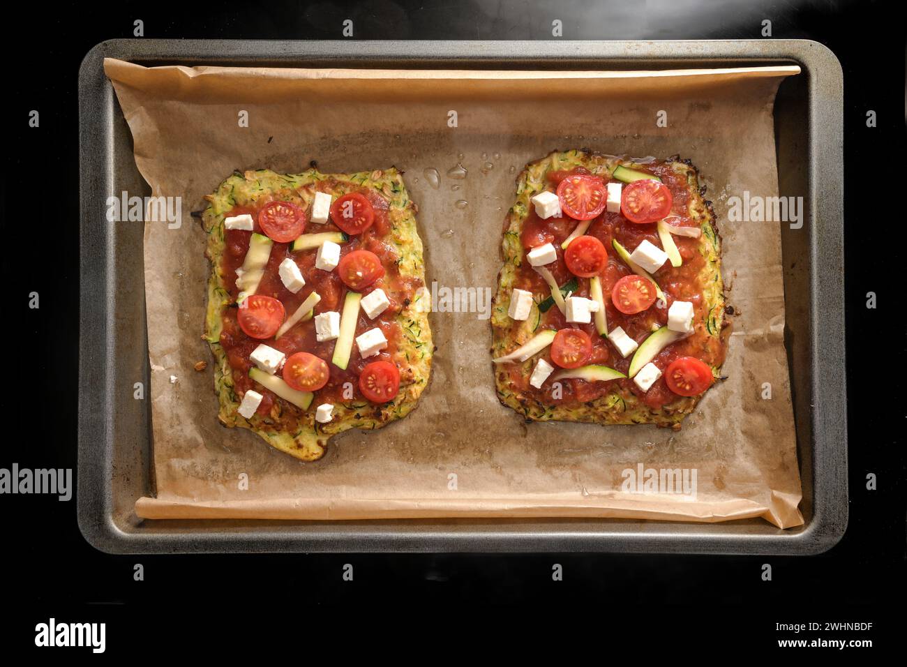 Vegetarian pizza pieces on a crust from zucchini and oat flakes, topped with tomatoes and feta cheese on a tray with baking pape Stock Photo