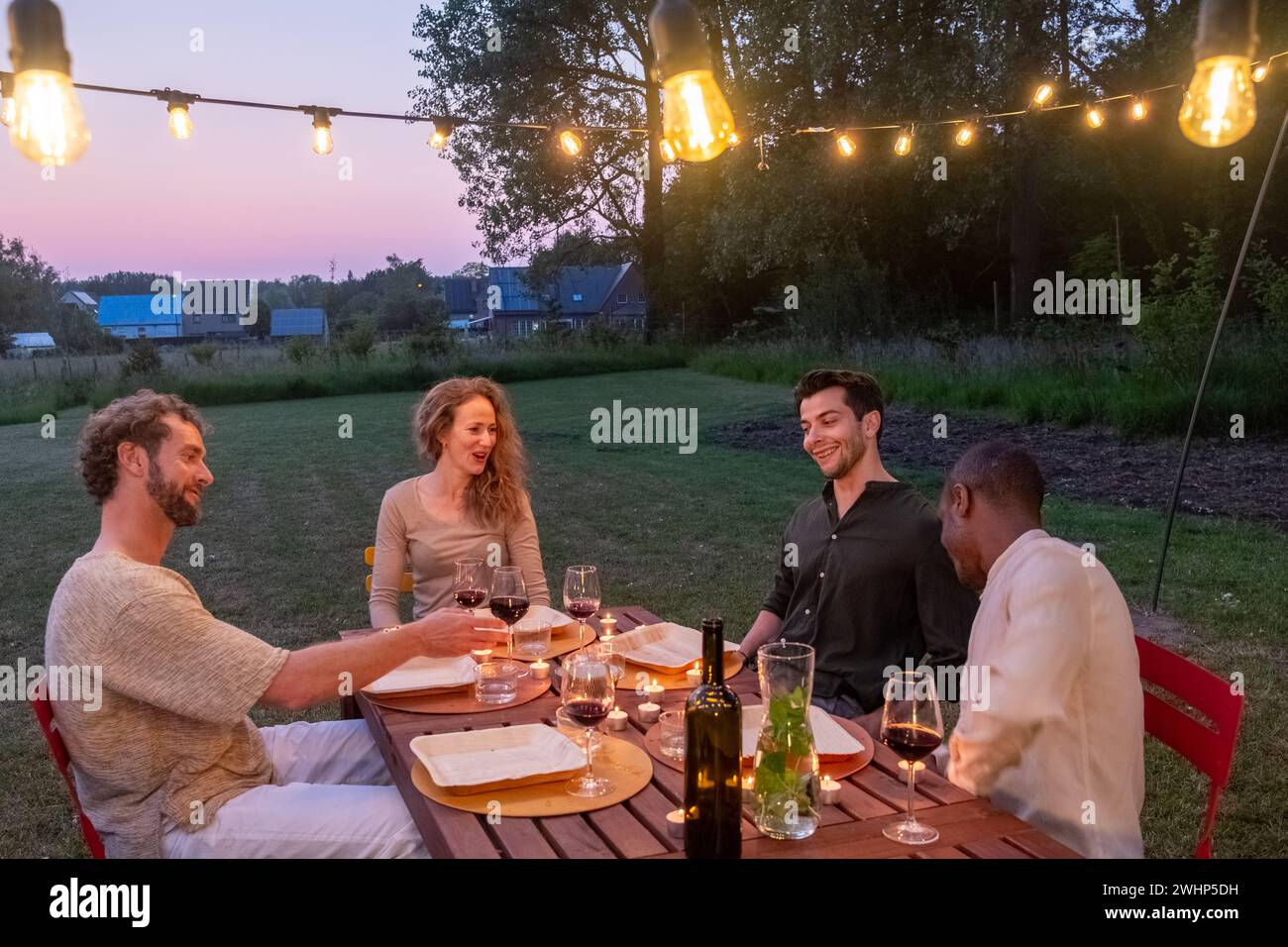 Multiracial group of millenial Friends Gathered at a Barbecue Dinner Table Outside a Beautiful Home with Lights at dusk Decorati Stock Photo