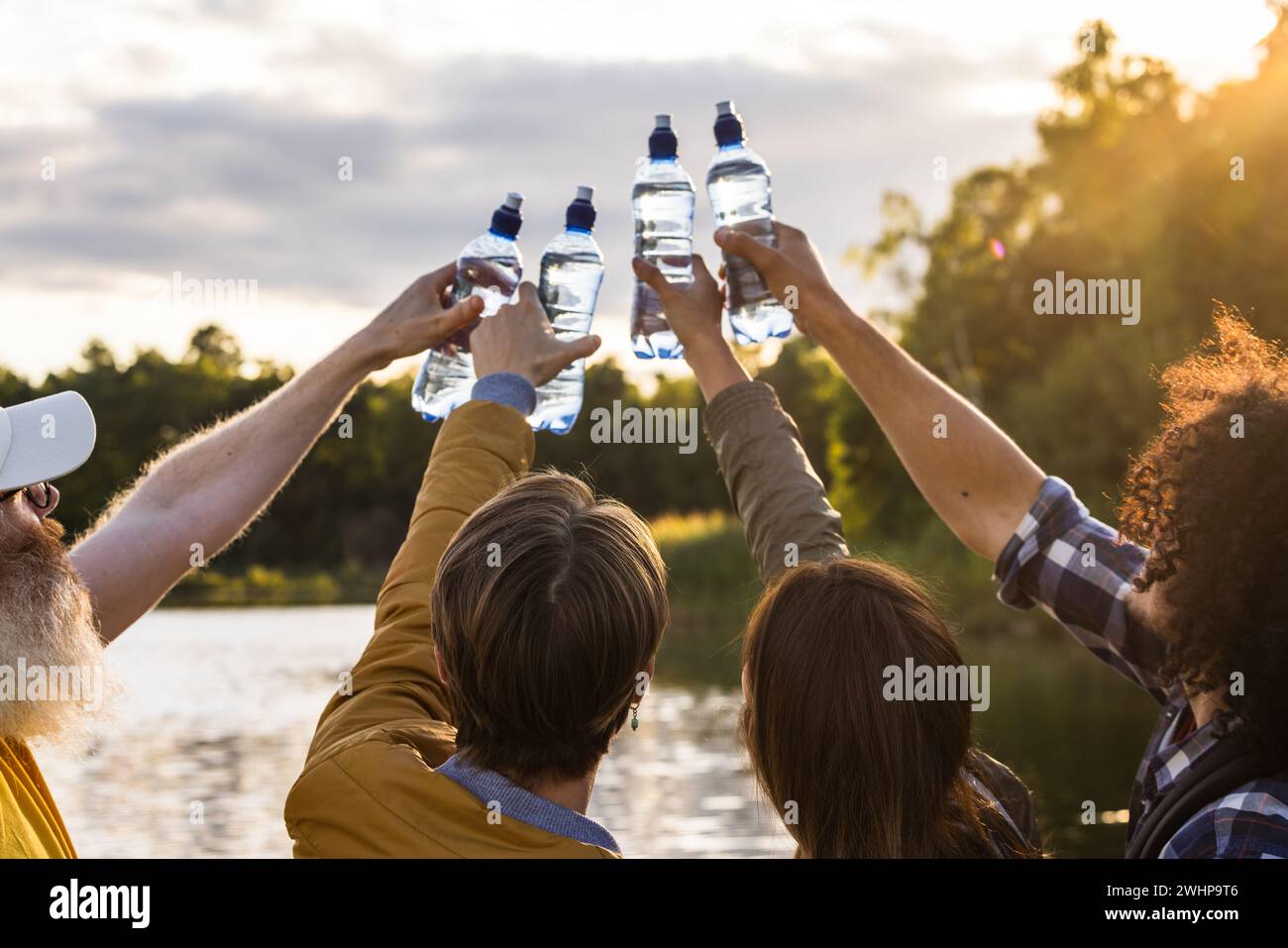 Four friends cheering with water bottles standing on the beach of a forest lake during beautiful sunset and celebrating life, re Stock Photo