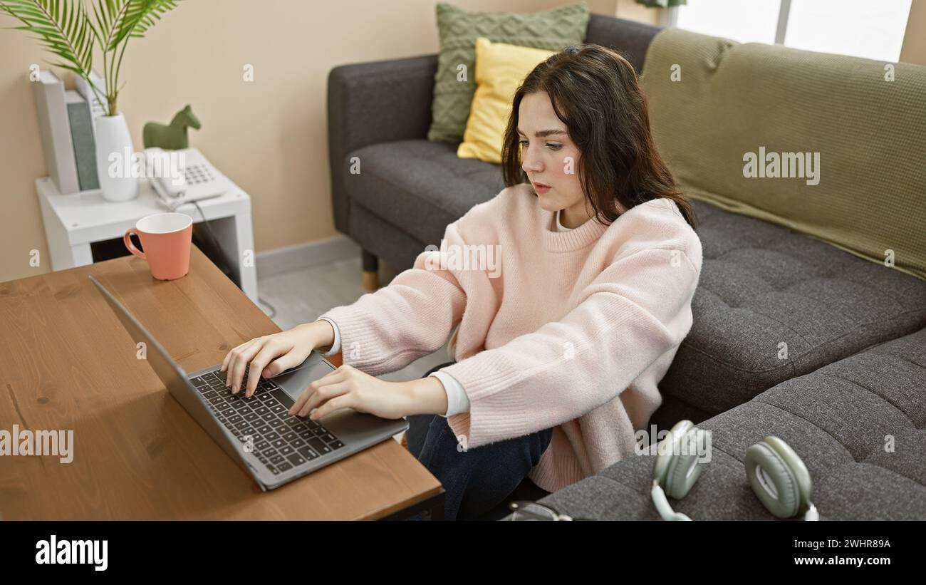 A young woman works on her laptop in a cozy living room, showcasing a ...
