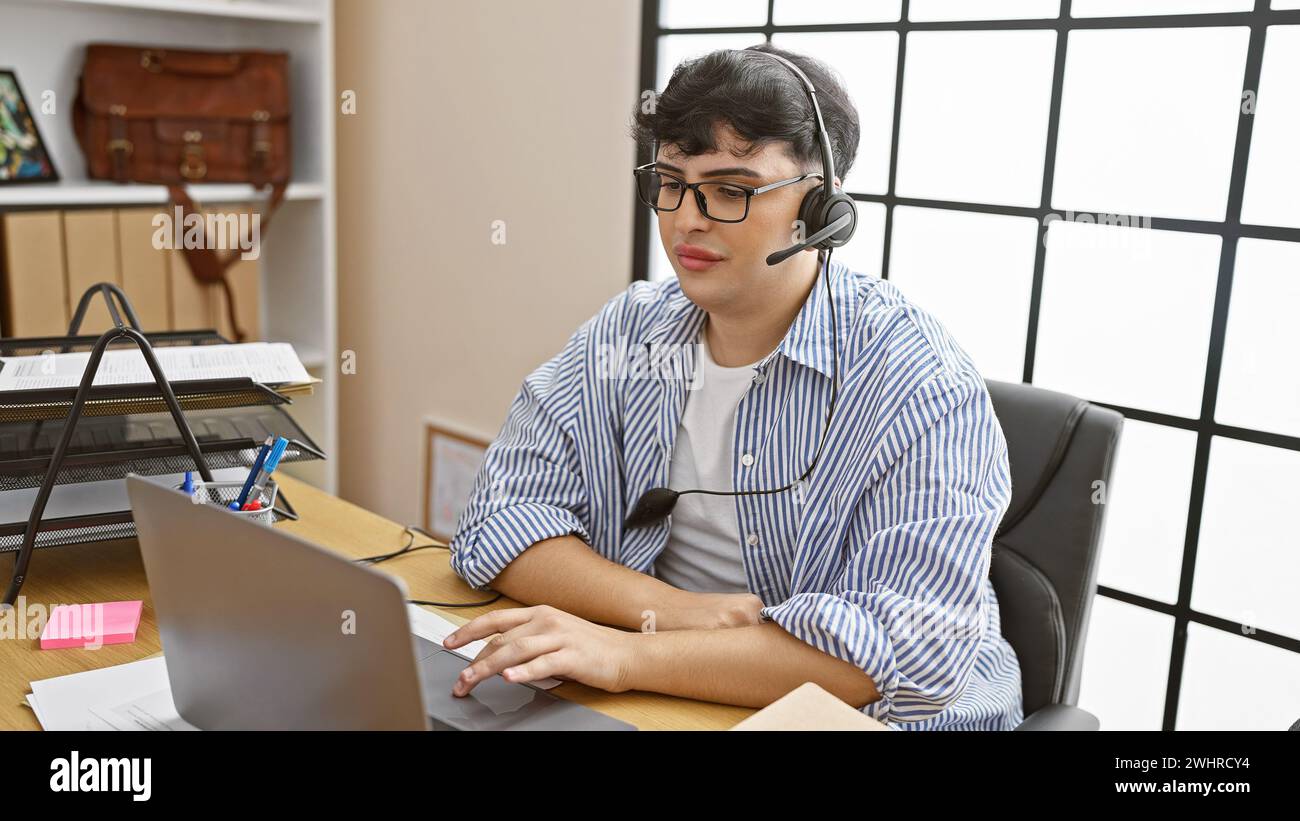 A young man in a striped shirt works diligently at his office desk, using a laptop and wearing a headset. Stock Photo